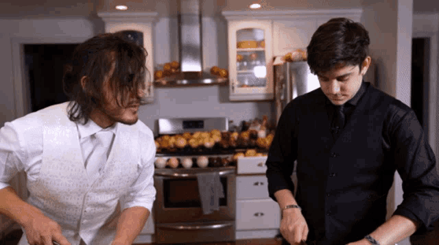 a man in a white vest stands next to a man in a black shirt in a kitchen