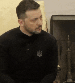 a man with a beard wears a black shirt with a symbol on the chest