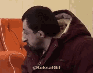a man in a red jacket is sitting on a couch with his nose in his mouth .