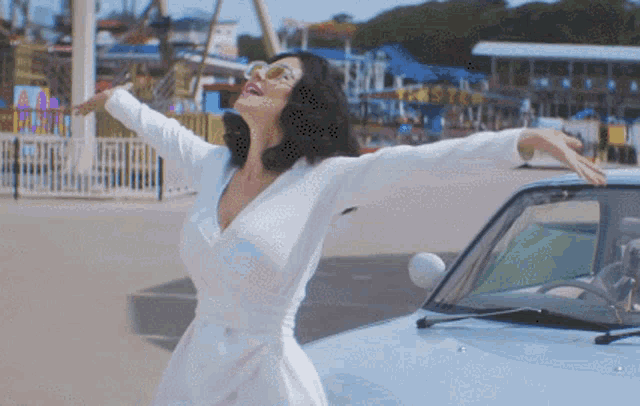 a woman in a white dress and sunglasses stands next to a car with her arms outstretched