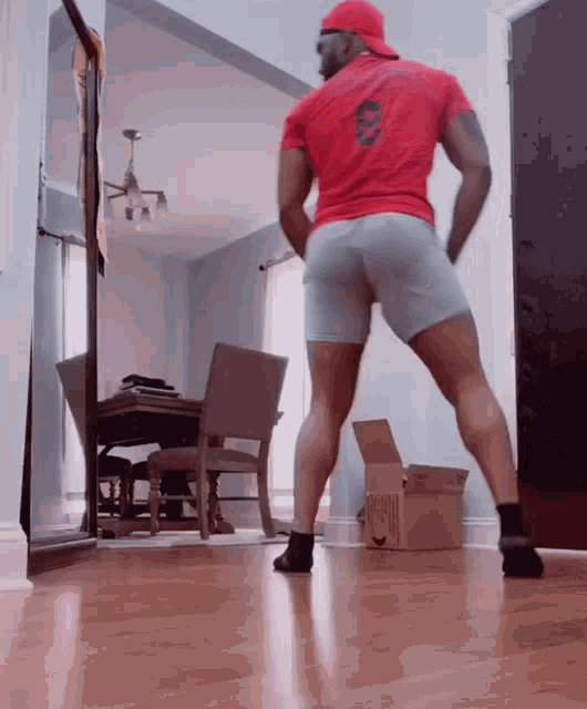 a man in a red shirt and grey shorts is standing on a wooden floor in a living room .