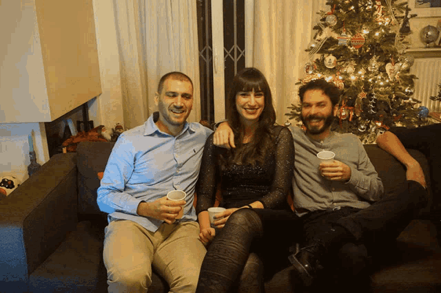 two men and a woman sitting on a couch with a christmas tree in the background