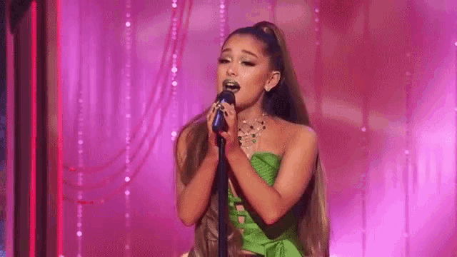 ariana grande is wearing a green top and singing into a microphone on stage .