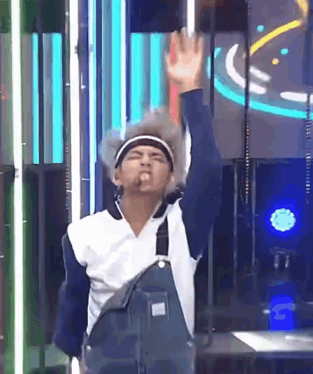 a man in overalls and a headband is standing on a stage with his hand up .
