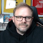 a man with glasses and a beard is wearing a black hoodie and sitting in a chair .