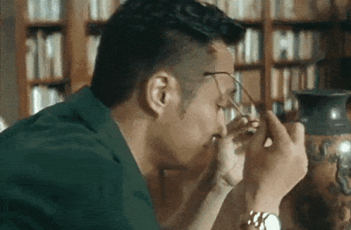a man wearing glasses looks at a vase in front of a bookshelf