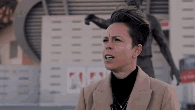a woman in a tan coat stands in front of a statue of a soccer player