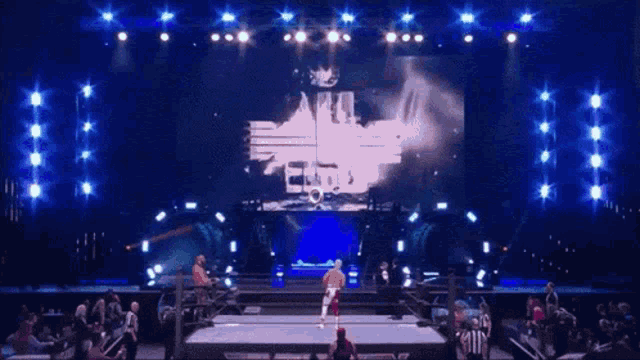 a man in a wrestling ring with a large screen behind him that says all