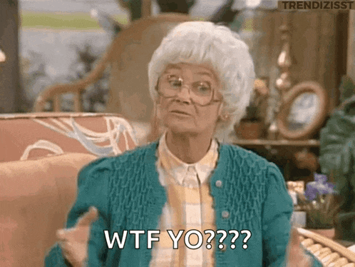 an elderly woman wearing glasses and a blue sweater is sitting on a couch and making a funny face .