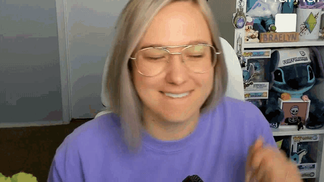 a woman wearing glasses and a purple shirt is smiling in front of a shelf with toys on it