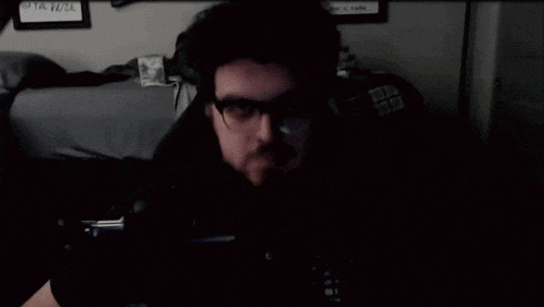 a man with glasses giving a thumbs up in front of a bed
