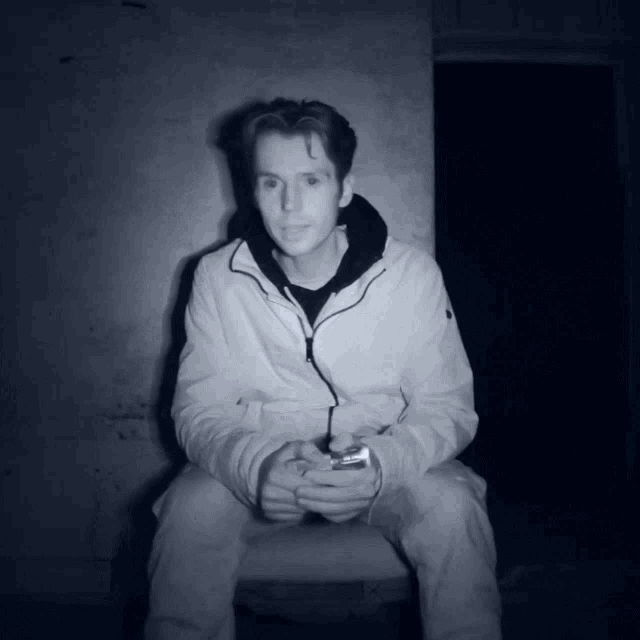 a man in a white jacket sits in a dark room with his hands folded