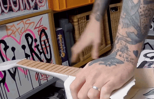 a person with a tattoo on their arm holds a guitar in front of a shelf with graffiti on it