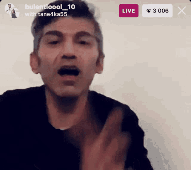 a man in a black shirt is talking in front of a white wall with a live button below him