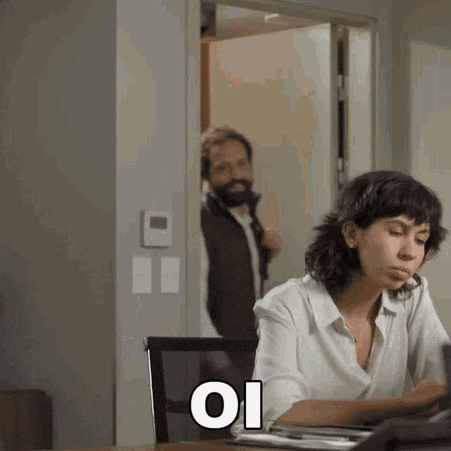 a woman sits at a desk with a man standing behind her and a sign that says oi on it
