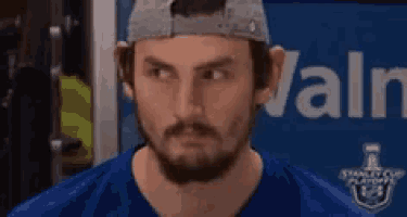 a man with a beard wearing a hat and a blue shirt is looking at the camera .