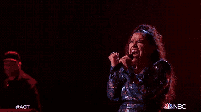 a woman is singing into a microphone with the nbc logo in the background