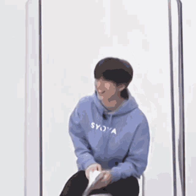 a man in a blue hoodie is sitting in front of a glass wall holding a book .