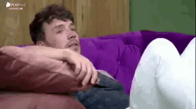 a man is laying on a purple couch with his legs crossed and looking at the camera .