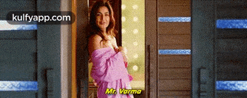a woman in a pink robe is standing in a doorway talking to mr. varma .