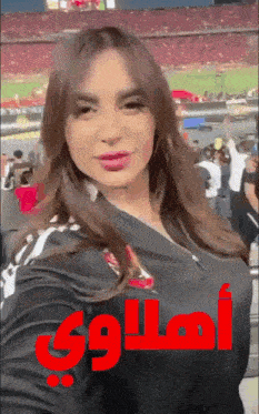 a woman taking a selfie in a stadium with arabic writing on the front of her shirt