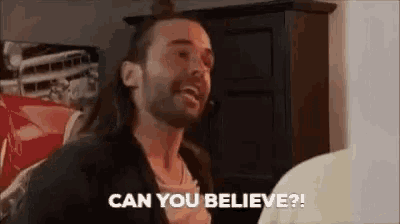 a man with long hair and a beard is talking to another man in a living room and says `` can you believe ? ''