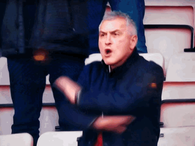 a man in a blue jacket is sitting in a stadium with his arms outstretched