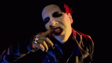 marilyn manson is wearing a leather jacket and a ring on his finger while making a face .
