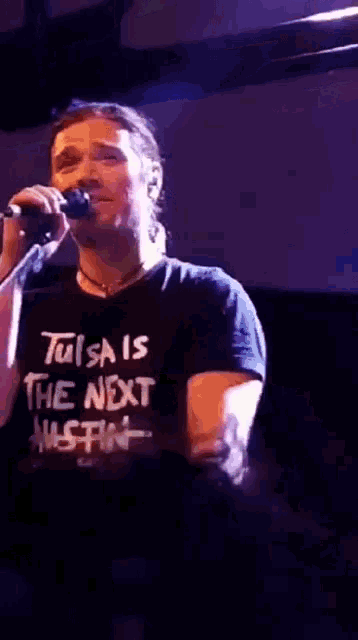a man singing into a microphone with a tulsa is the next austin shirt on