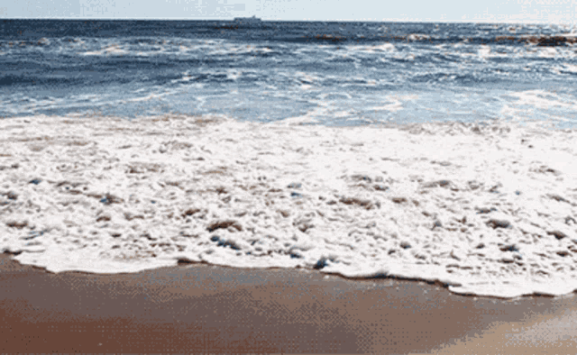 waves crashing on a sandy beach with a large ship in the distance