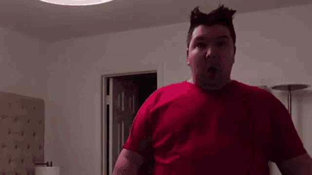 a man in a red shirt is standing in front of a bed in a bedroom .
