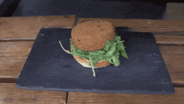 a person is putting lettuce on a hamburger on a slate cutting board