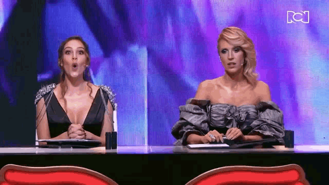 two women are sitting at a table with a purple background and the letter c on the bottom