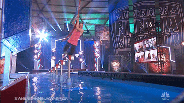 a man is hanging from a rope in a pool with a sign that says american ninja warrior in the background