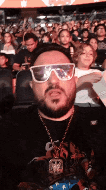 a man wearing sunglasses and a necklace with the letter w on it sits in a crowd