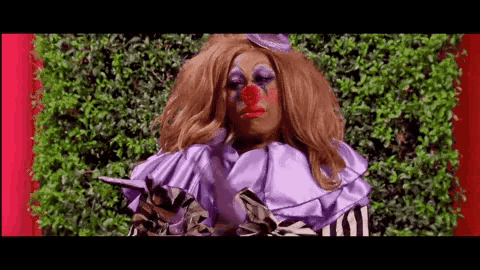 a woman in a clown costume is standing in front of a green bush .