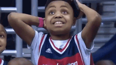 a young boy in a washington wizards jersey is sitting in the stands with his hands on his head .