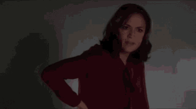 a woman in a red sweater is standing in a dark room with her hand on her hips .