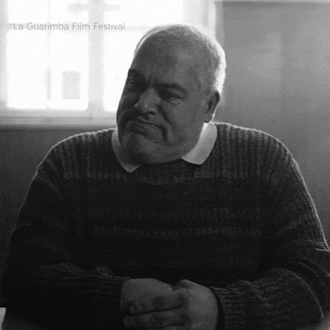 a man in a sweater sits at a table with his hands folded in front of a la guarimba film festival sign
