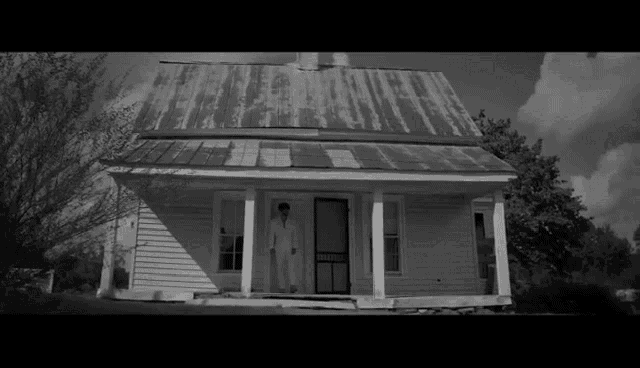 a black and white photo of a man standing on the porch of an old white house