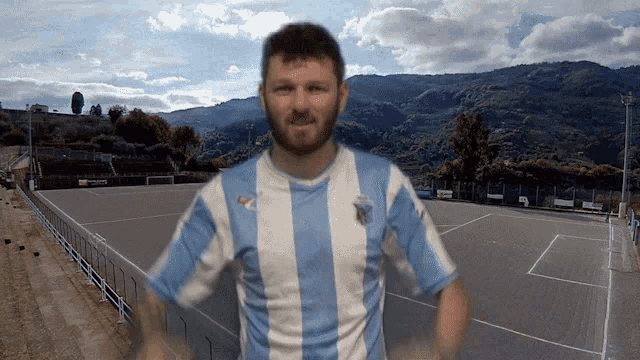 a man with a beard wearing a blue and white striped shirt with a soccer field behind him