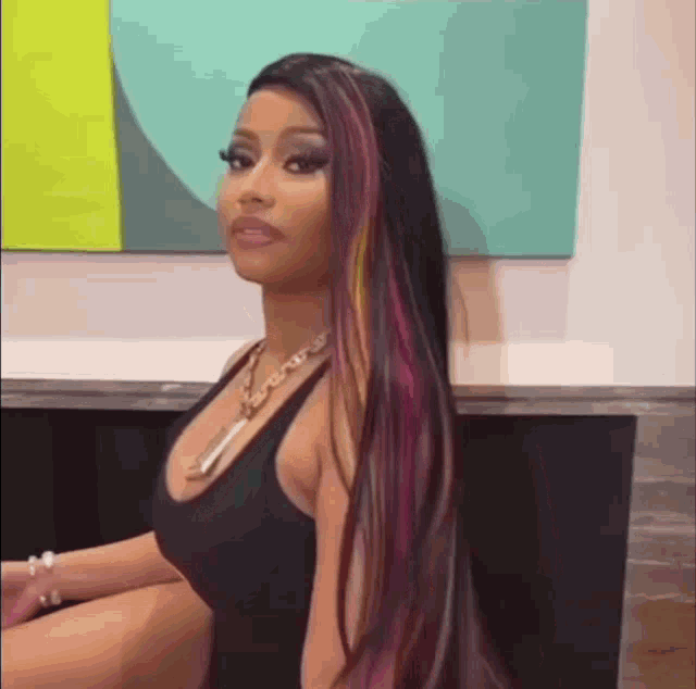 nicki minaj is wearing a black tank top and a necklace while sitting in front of a painting .