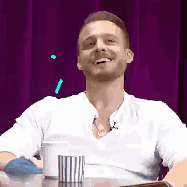 a man in a white shirt is sitting at a table with a cup of coffee and smiling .