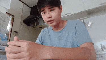 a young man in a blue shirt is sitting in a kitchen with his hands folded in front of him