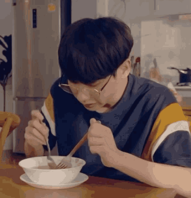 a young man wearing glasses is sitting at a table eating a bowl of soup with a fork