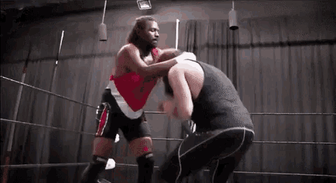 two men are wrestling in a ring and one is wearing a red tank top .