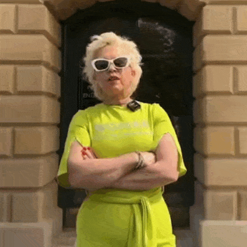 a woman in a neon yellow dress and white sunglasses is standing with her arms crossed in front of a building .