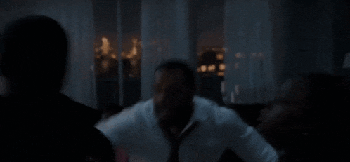 a man in a white shirt is sitting in a dark room looking out a window