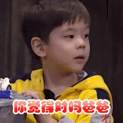 a young boy in a yellow and blue jacket is sitting in front of a trash can .