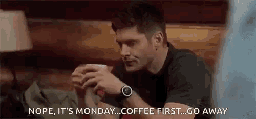 a man is sitting at a table holding a cup of coffee and talking to another man .